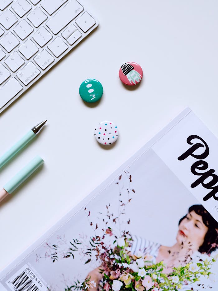 Flat lay of a contemporary desk setup with keyboard, pens, buttons, and a magazine on a white background.